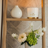 A lit Melissa Wilkes England scented candle which is used as a thing of beauty together with a small vase and beautiful small fresh flower arrangement of rose and sweet pea. The gold lid is sat underneath the lit candle acting as a coaster whilst still showing casing some of the gorgeous gold to compliment the gold branding on a background of white bone china. 