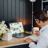 This is a photograph of Melissa at her wooden top desk with her favourite silver fountain pen creating her to do list for the week ahead in her paper diary.  Lit on the desk is her citrus scented candle Pomelo Bitters. The flame is slightly out of focus creating a warm glow in the picture.  There is the candles pristine white gift box which is hand wrapped in England. There is also a small vase brimming with white sweet peas and roses.  Melissa always has fresh flowers  at her desk. 